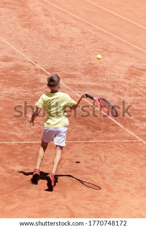 Child boy plays tennis on clay court. Young male tennis player hits the ball in flight in action. Kids tennis concept. Sports action frame. Motion. Vertical banner with copy space