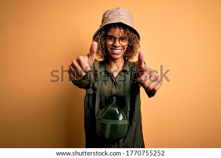 Young african american tourist woman on vacation wearing explorer hat and water canteen approving doing positive gesture with hand, thumbs up smiling and happy for success. Winner gesture.