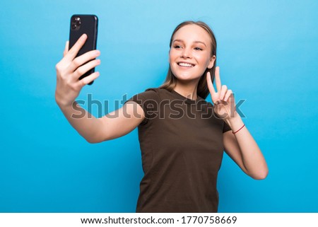 Cheerful young woman raising hand in hello gesture and smiling happily while making video call with her mobile phone, isolated over blue background
