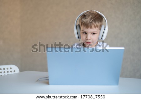 Cute little boy is watching cartoons or a movie on a laptop. Caucasian child sits at a table at home and listens to music on headphones.
