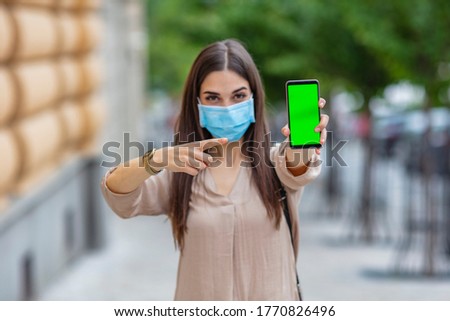 Young Woman In Medical Mask Pointing At Smartphone With green Screen in the street. Woman wearing coronavirus protection mask showing smartphone blank screen