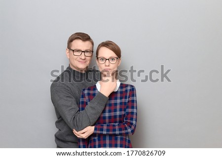 Portrait of funny weird couple, woman with serious expression, man is making goofy ridiculous faces, having fun, fooling around behind her, both are standing over gray background. Relationship concept