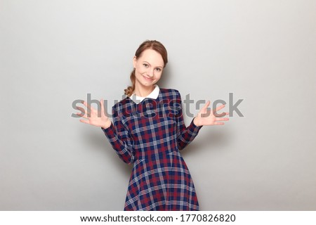 Studio portrait of happy carefree joyful girl wearing checkered dress, smiling cheerfully, raising hands up, dancing, celebrating victory or success, being in good mood, standing over gray background