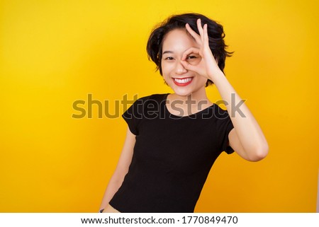 Beautiful young asian woman with happy face smiling doing ok sign with hand on eye looking through finger. Wearing casual clothes and standing.