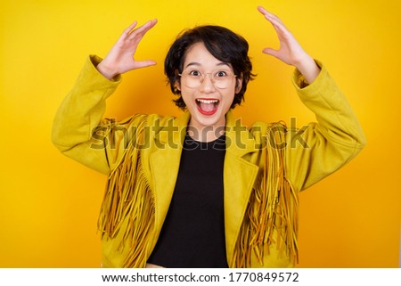 Portrait of beautiful asian female looks with excitement at camera, keeps hands raised over head, notices something unexpected, isolated over gray wall. Lovely woman reacts on sudden news.
