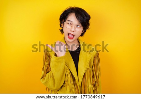 Omg concept. Stupefied asian female with surprised expression, opens eyes and mouth widely, points aside with thumb, shows something strange on gray background. Advertisement concept.