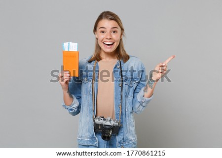 Excited girl in denim clothes with photo camera isolated on gray wall background. Passenger traveling abroad on weekends getaway. Air flight concept. Hold passport tickets pointing index finger aside