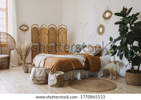Comfortable apartment in bohemian style interior with hygge bedroom, pillow and bedspread on bed, bamboo dressing screen, home decor, dry plants in vase, wicker basket, houseplant on floor Royalty-Free Stock Photo #1770865523