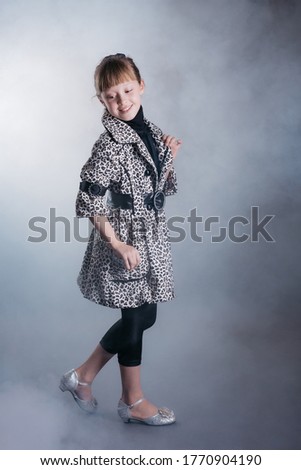 Little girl in leopard print coat, black leggings and silver shoes against a dark background in smoke