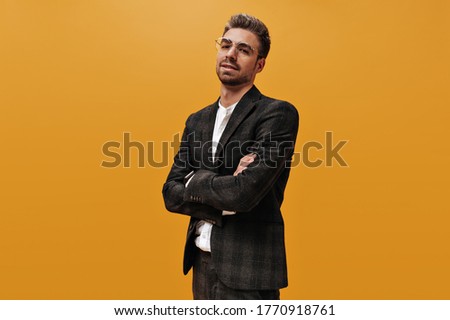 Cool self-confident man in checkered jacket and white t-shirt looks into camera. Bearded guy crosses arms and poses on orange background.