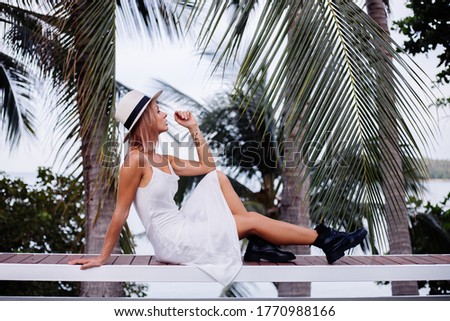 Fashion gentle portrait of elegant stylish woman in white sild long dress, big black boots and classic viscose white hat, tropical exotic background, outdoor shot.