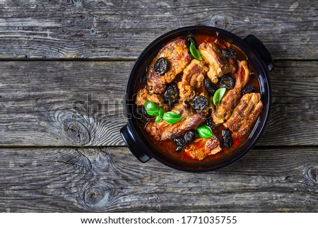 Stewed short pork ribs with dried plums or prunes, pork broth, onion, and garlic, decorated with fresh basil, served on a black baking dish on an old barn wood background, copy space