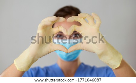 Young girl in a blue medical attire looking at the camera through a heart shape formed with gloved fingers. Picture of a close-up heart symbol.