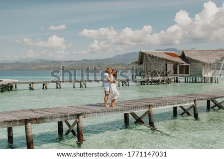 Couple on the beach at tropical resort. Travel honeymoon concept. Happy loving couple in white clothes hug each other while stand on wooden jetty