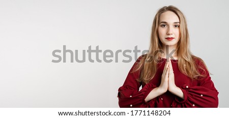 Please, help me. Emotive pleased European girl with long blonde hair, keeps hands in praying gesture, beggs for giving good advice, dressed in red blouse. Isolated over white background.