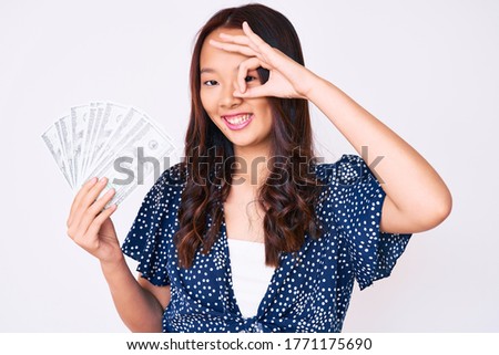 Young beautiful chinese girl holding dollars smiling happy doing ok sign with hand on eye looking through fingers 
