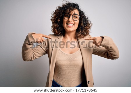 Young curly arab business woman wearing elegant jacket and glasses over white background looking confident with smile on face, pointing oneself with fingers proud and happy.