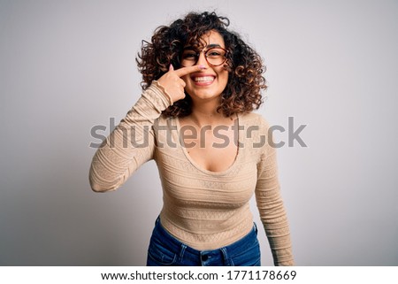 Young beautiful curly arab woman wearing casual t-shirt and glasses over white background Pointing with hand finger to face and nose, smiling cheerful. Beauty concept
