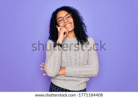 Young african american woman wearing casual sweater and glasses over purple background looking confident at the camera smiling with crossed arms and hand raised on chin. Thinking positive.