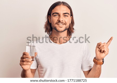Young handsome man drinking glass of water smiling happy pointing with hand and finger to the side 