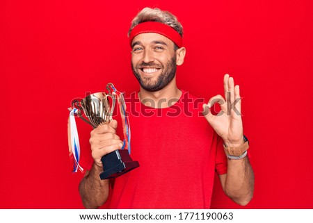 Young handsome blond sportsman with beard winning trophy over isolated red background doing ok sign with fingers, smiling friendly gesturing excellent symbol
