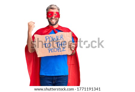 Young blond man wearing super hero custome holding power to the people cardboard banner screaming proud, celebrating victory and success very excited with raised arms 