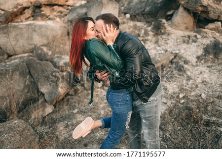 Beautiful stylish young couple in love in leather jackets hugging and passionate kissing in the summer on a sand quarry. Woman with red hair, man with a watch on his hands.