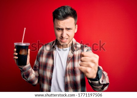 Young handsome caucasian man drinking a fresh soda refreshment over red background annoyed and frustrated shouting with anger, crazy and yelling with raised hand, anger concept