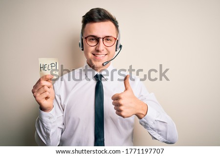 Young call center operator man wearing headset holding paper note with help word happy with big smile doing ok sign, thumb up with fingers, excellent sign