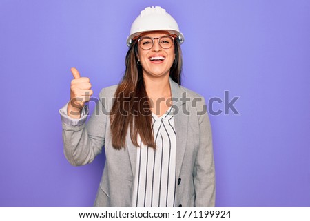 Professional woman engineer wearing industrial safety helmet over pruple background doing happy thumbs up gesture with hand. Approving expression looking at the camera showing success.