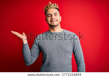 Young handsome man wearing golden crown of prince over isolated red background smiling cheerful presenting and pointing with palm of hand looking at the camera.