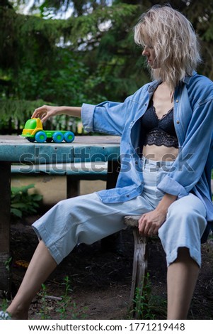 A girl in a denim suit sits on a bench at a table outside in the garden and rolls a small toy car. Adult childhood memories, boredom