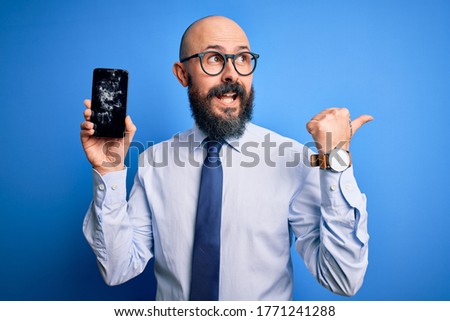 Handsome bald business man with beard holding broken smartphone showing cracked screen pointing and showing with thumb up to the side with happy face smiling