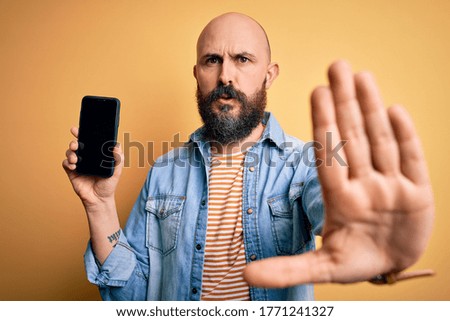 Handsome bald man with beard holding smartphone showing screen over yellow background with open hand doing stop sign with serious and confident expression, defense gesture