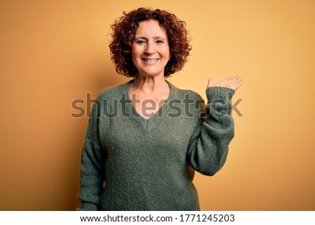 Middle age beautiful curly hair woman wearing casual sweater over isolated yellow background smiling cheerful presenting and pointing with palm of hand looking at the camera.