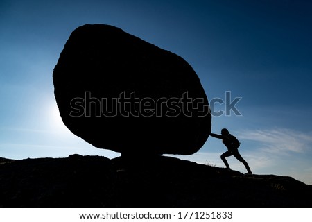 Silhouette of woman with backpack on background blue sky pushing huge rock