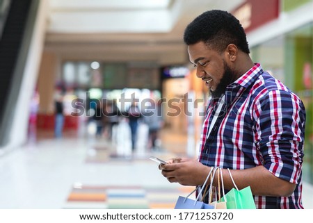 App With Discounts. Smiling Black Man With Shopping Bags Using Smartphone In Mall, Checking Sale Deals, Side View With Free Space Royalty-Free Stock Photo #1771253720