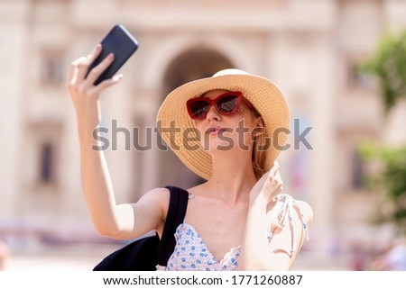 Portrait shot of happy young woman wearing straw hat and using mobile phone while making selfie on the street. 