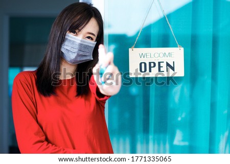 Asian woman shop owner smiling hanging label 'Welcom we are open' and showing thump up. She open her shop after pandemic of coronavirus outbreak