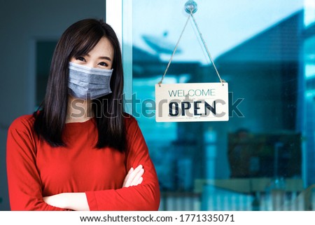 Asian woman shop owner smiling hanging label 'Welcom we are open' and showing thump up. She open her shop after pandemic of coronavirus outbreak