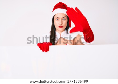 Young woman wearing santa claus costume holding blank empty banner with open hand doing stop sign with serious and confident expression, defense gesture 