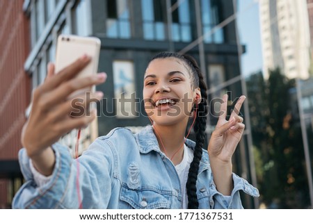 Beautiful African American woman with stylish hairstyle taking selfie, showing victory sign, standing outdoors. Young emotional blogger influencer using mobile phone, streaming video on urban street