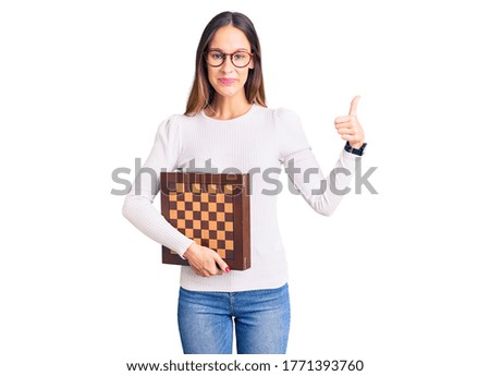 Beautiful brunette young woman holding chess smiling happy and positive, thumb up doing excellent and approval sign 