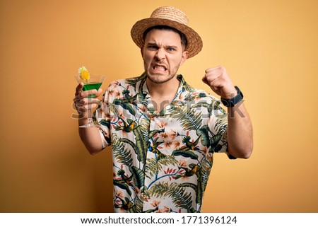 Young man on vacation wearing summer hat drinking a party cocktail over yellow background annoyed and frustrated shouting with anger, crazy and yelling with raised hand, anger concept