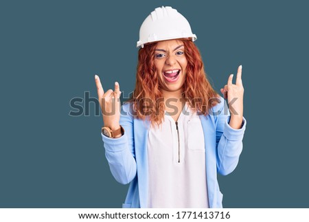 Young latin woman wearing architect hardhat shouting with crazy expression doing rock symbol with hands up. music star. heavy concept. 