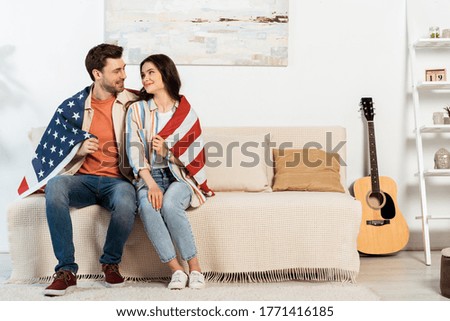 Young couple wrapping in american flag smiling at each other on couch