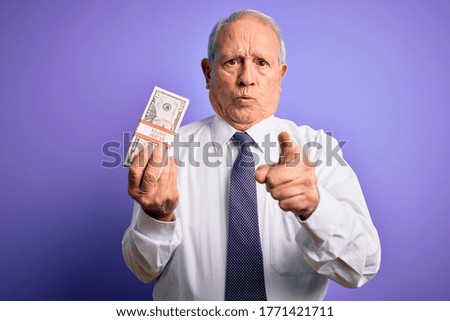 Senior grey haired man holding bunch of fifty dollars banknotes over purple background pointing with finger to the camera and to you, hand sign, positive and confident gesture from the front