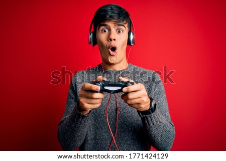 Young handsome gamer man playing video game using joystick and headphones scared in shock with a surprise face, afraid and excited with fear expression