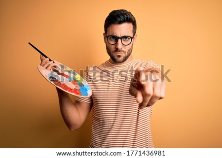 Young handsome artist man with beard painting using bursh and palette with colors pointing with finger to the camera and to you, hand sign, positive and confident gesture from the front