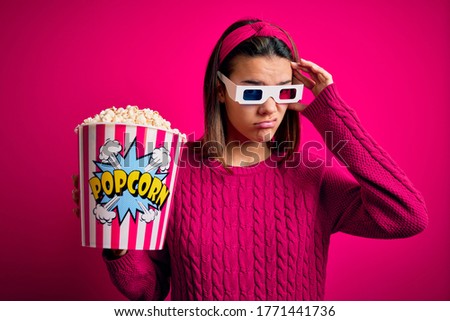 Young beautiful girl watching movie using 3d glasses eating box with popcorns worried and stressed about a problem with hand on forehead, nervous and anxious for crisis